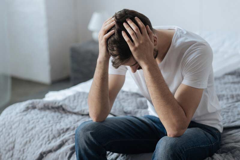 Mann sitzend auf Bettrand, hält seinen Kopf verstört, fragt sich, was die Ursache seiner Probleme beim Sex ist